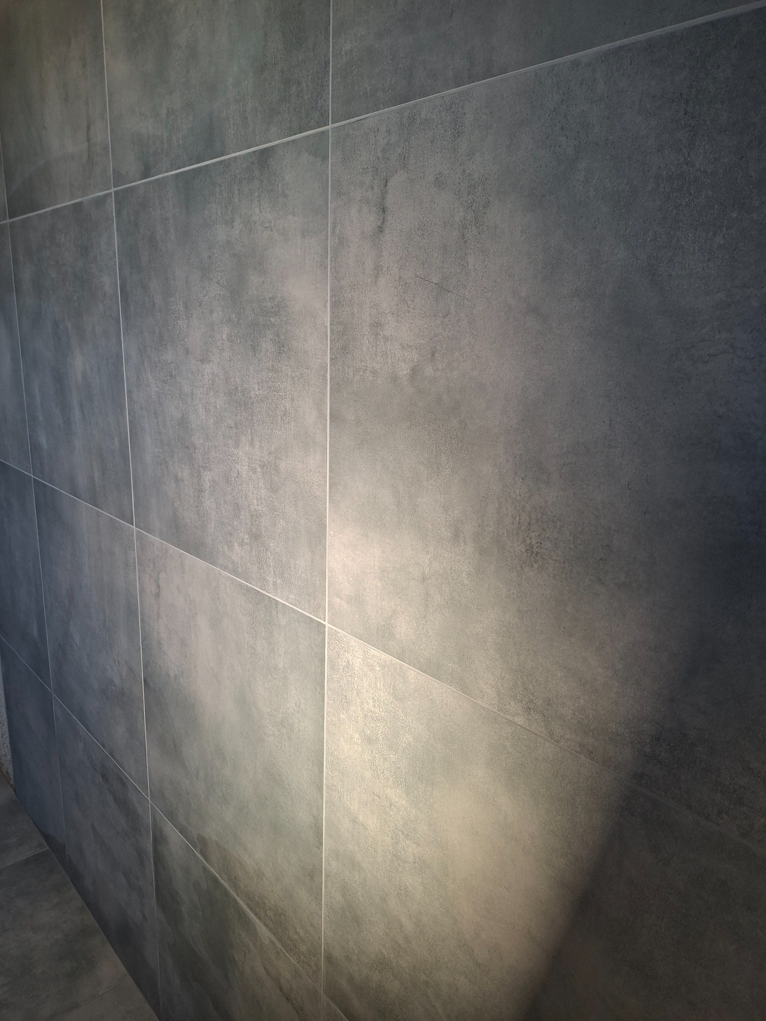 Ściana wyłożona szarymi, matowymi płytkami o dużym formacie, z delikatną fakturą betonu, oświetlona z boku, tworząca minimalistyczny i nowoczesny efekt.