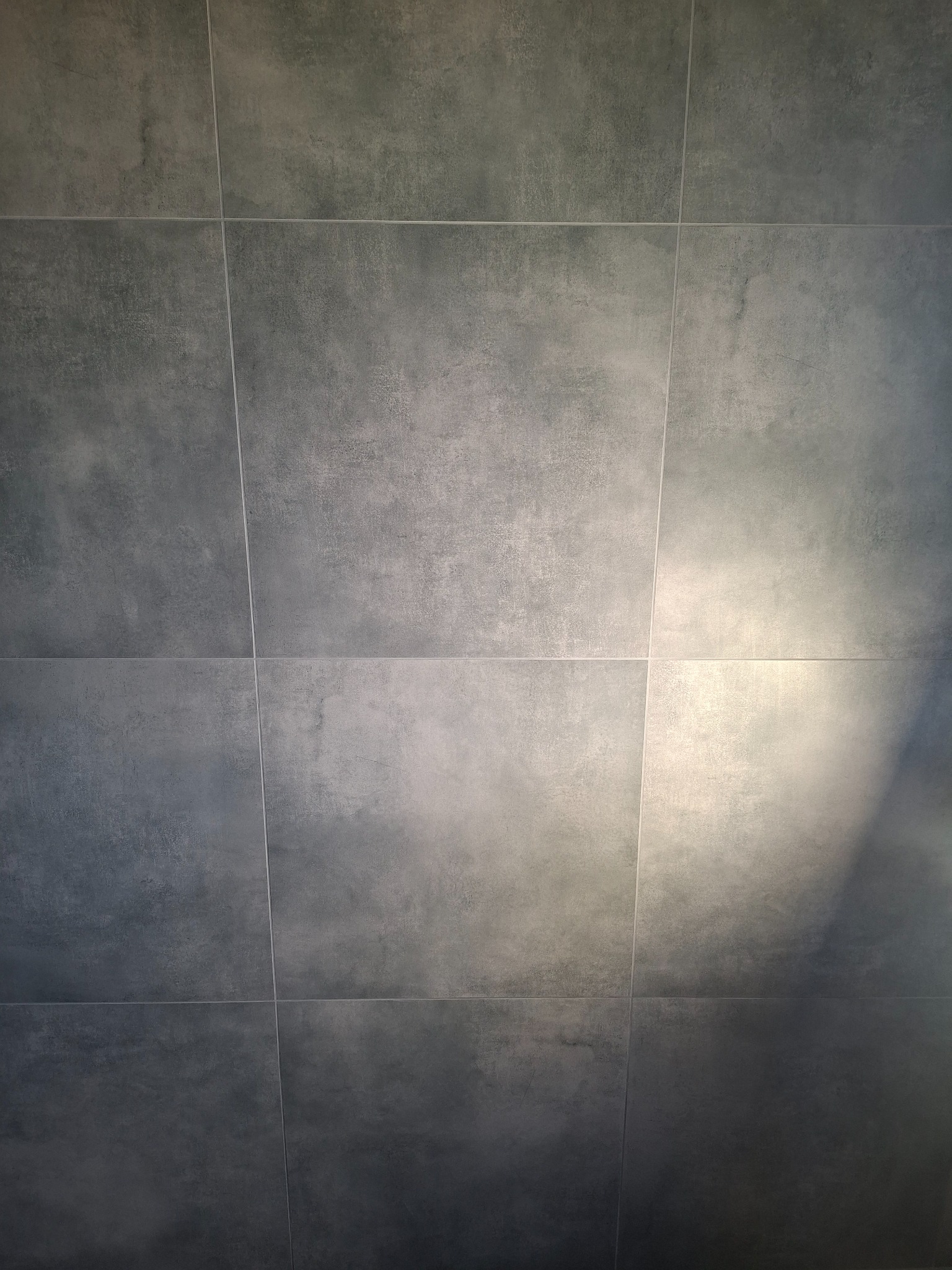 Szare, matowe płytki ceramiczne ułożone w regularną siatkę, z delikatnym gradientem światła, tworzące nowoczesną powierzchnię ściany lub podłogi.