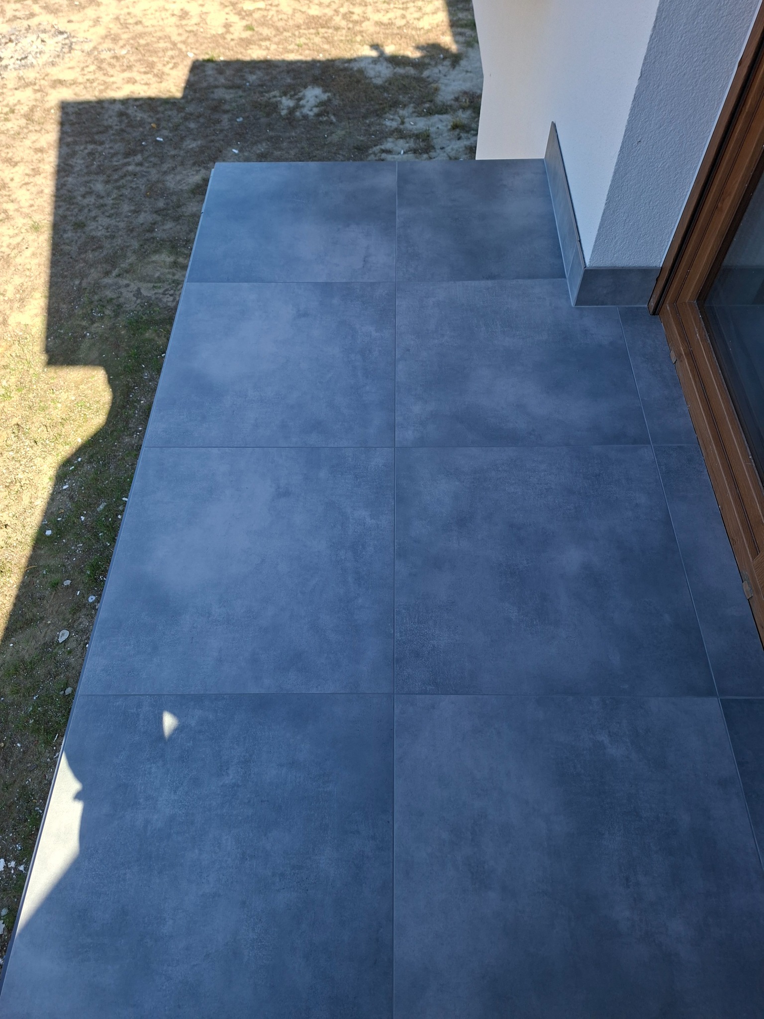 Szare, matowe płytki gresowe ułożone na zewnątrz, tworzące taras przy budynku z drewnianą ramą okienną. Widoczne fugi i krawędź tarasu.