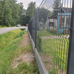 Nowoczesne, grafitowe ogrodzenie z paneli z podmurówką betonową, widok z boku na tle zieleni, drogi i budynku.