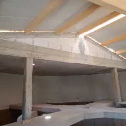 Wnętrze surowego budynku w trakcie budowy z betonowymi słupami, stropem i podłogą, drewnianą konstrukcją dachu i ścianami z bloczków betonowych.