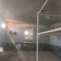 Grzechu Grzegorz Jasiński - Wnętrze w trakcie prac wykończeniowych: ściany pokryte świeżym tynkiem, zabezpieczona folią podłoga, widoczne rusztowanie i wiadro z zaprawą.