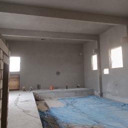 Grzechu Grzegorz Jasiński - Surowe wnętrze budynku w trakcie prac wykończeniowych, ściany przygotowane pod wykończenie, zabezpieczona podłoga, widoczne otwory okienne i fragment murowanej ściany.