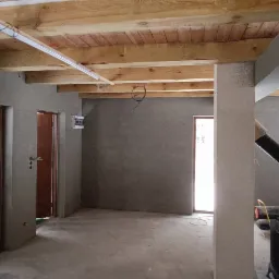 Wnętrze w stanie surowym: ściany pokryte szarym tynkiem, drewniany strop z widocznymi belkami, dwie drewniane ościeżnice drzwiowe, widoczne przewody elektryczne, kolumna z surowym wykończeniem.