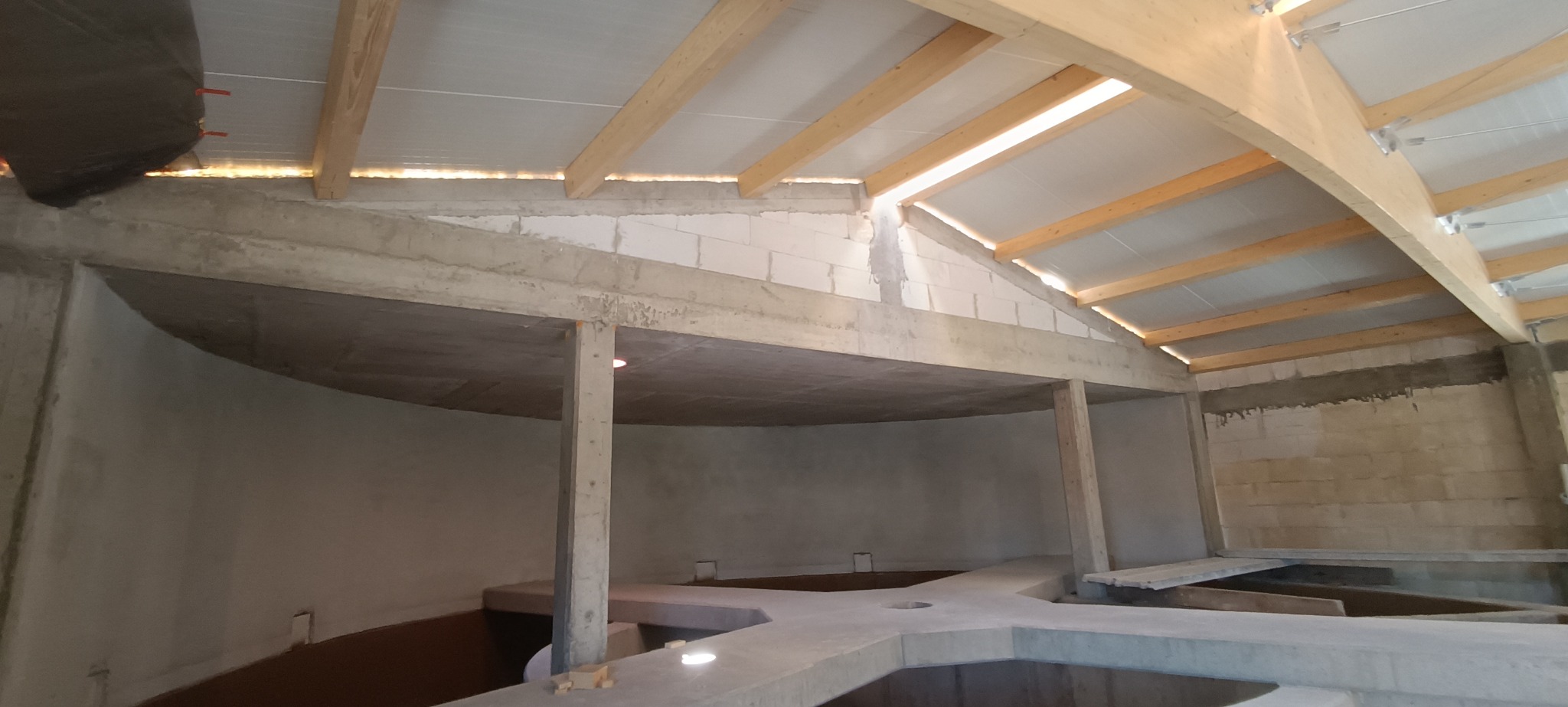 Wnętrze surowego budynku w trakcie budowy z betonowymi słupami, stropem i podłogą, drewnianą konstrukcją dachu i ścianami z bloczków betonowych.