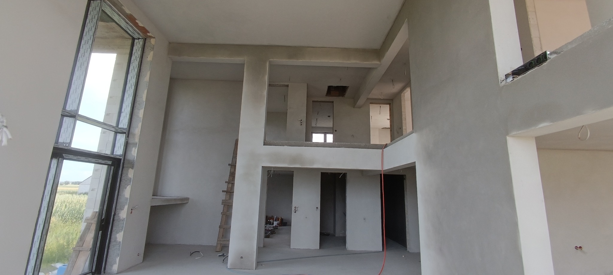 Wnętrze dwukondygnacyjnego domu w trakcie prac wykończeniowych, widoczne ściany po nałożeniu warstwy wykończeniowej, surowe okno PCV z widokiem na zewnątrz, drewniana drabina oparta o ścianę, kable...