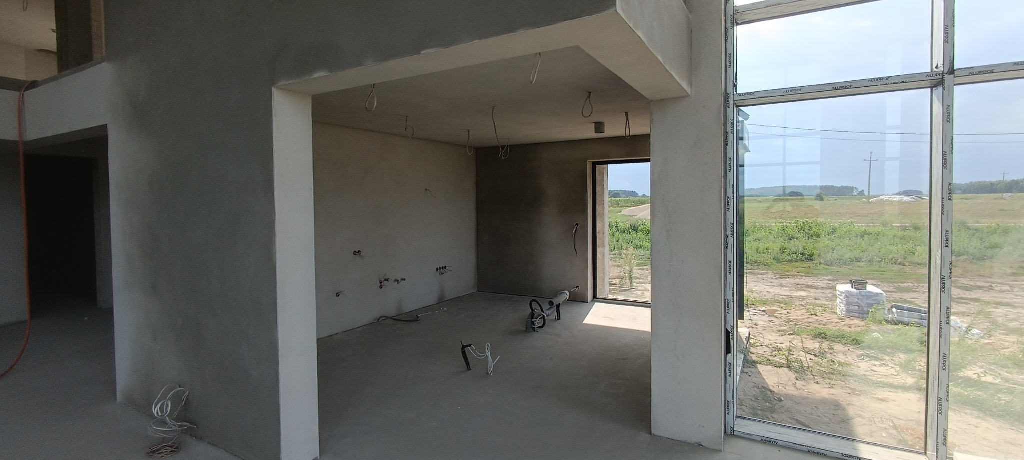 Surowe wnętrze w trakcie wykańczania, ściany pokryte szarym tynkiem, widoczne przewody elektryczne, duże okno z widokiem na zewnątrz, worki materiałów budowlanych poza budynkiem.