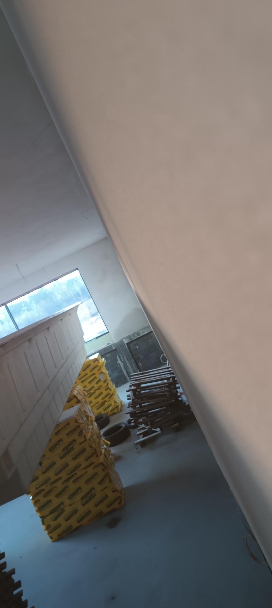 Świeżo wykończona ściana z gładzią, widoczne materiały budowlane i narzędzia w tle, okno z widokiem na zewnątrz.