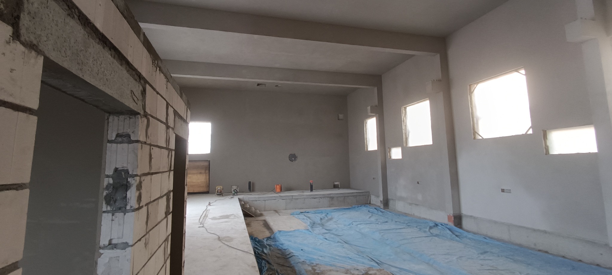 Surowe wnętrze budynku w trakcie prac wykończeniowych, ściany przygotowane pod wykończenie, zabezpieczona podłoga, widoczne otwory okienne i fragment murowanej ściany.