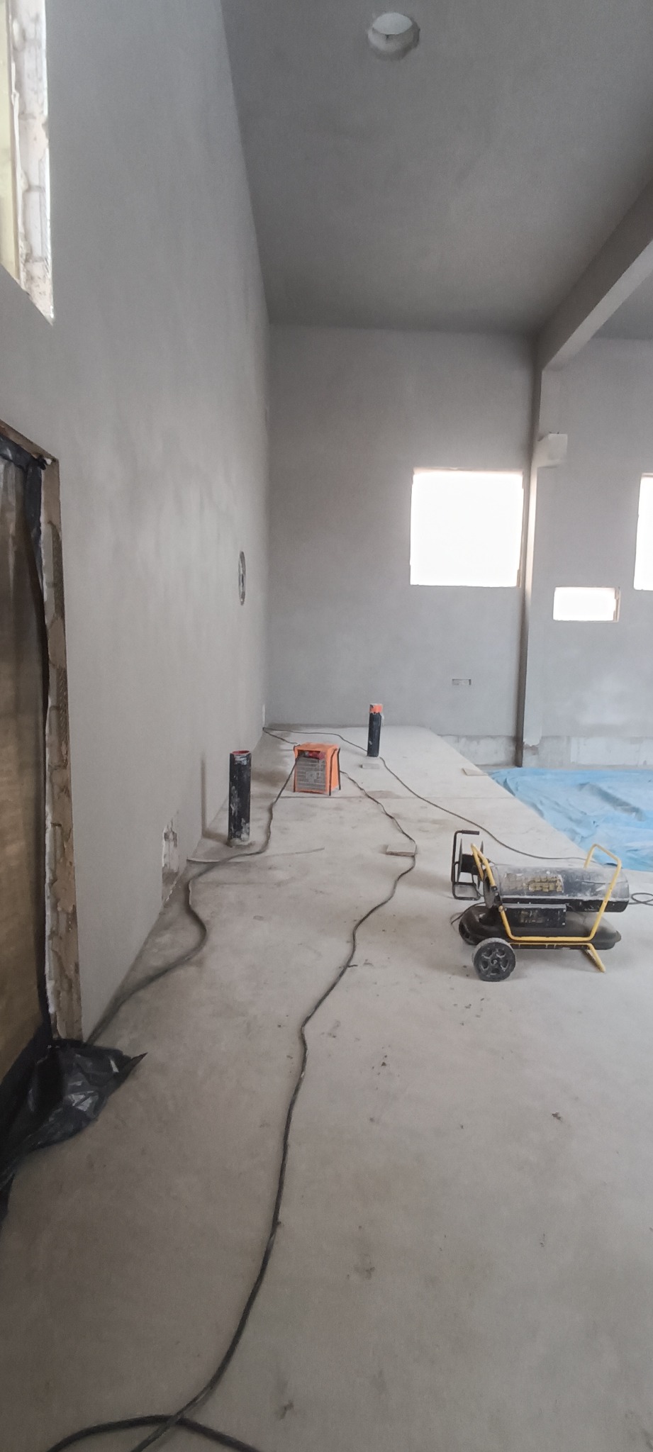 Świeżo otynkowane ściany w surowym pomieszczeniu z widocznym nagrzewnicą, przewodami i elementami instalacji elektrycznej przed wykończeniem podłogi.