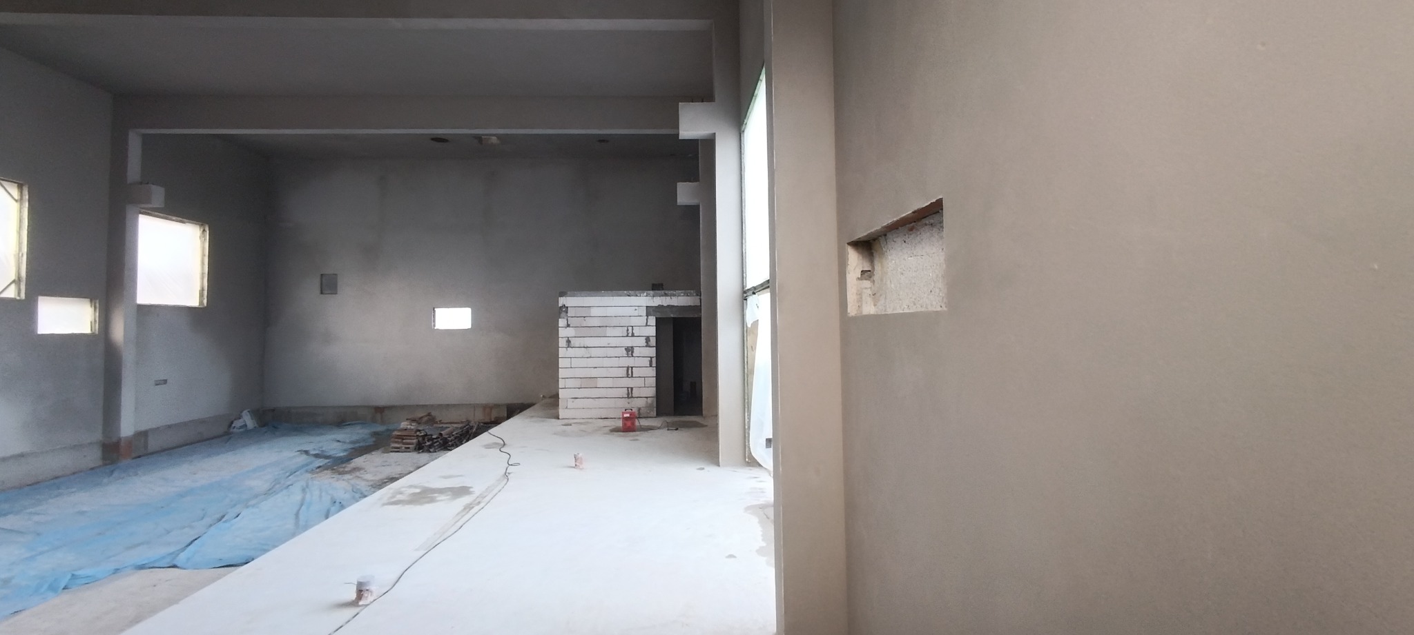Wnętrze w trakcie prac wykończeniowych: ściany pokryte szarym tynkiem, widoczny fragment murowanej ściany z bloczków, podłoga częściowo zabezpieczona folią ochronną, małe okno w głębi pomieszczenia.