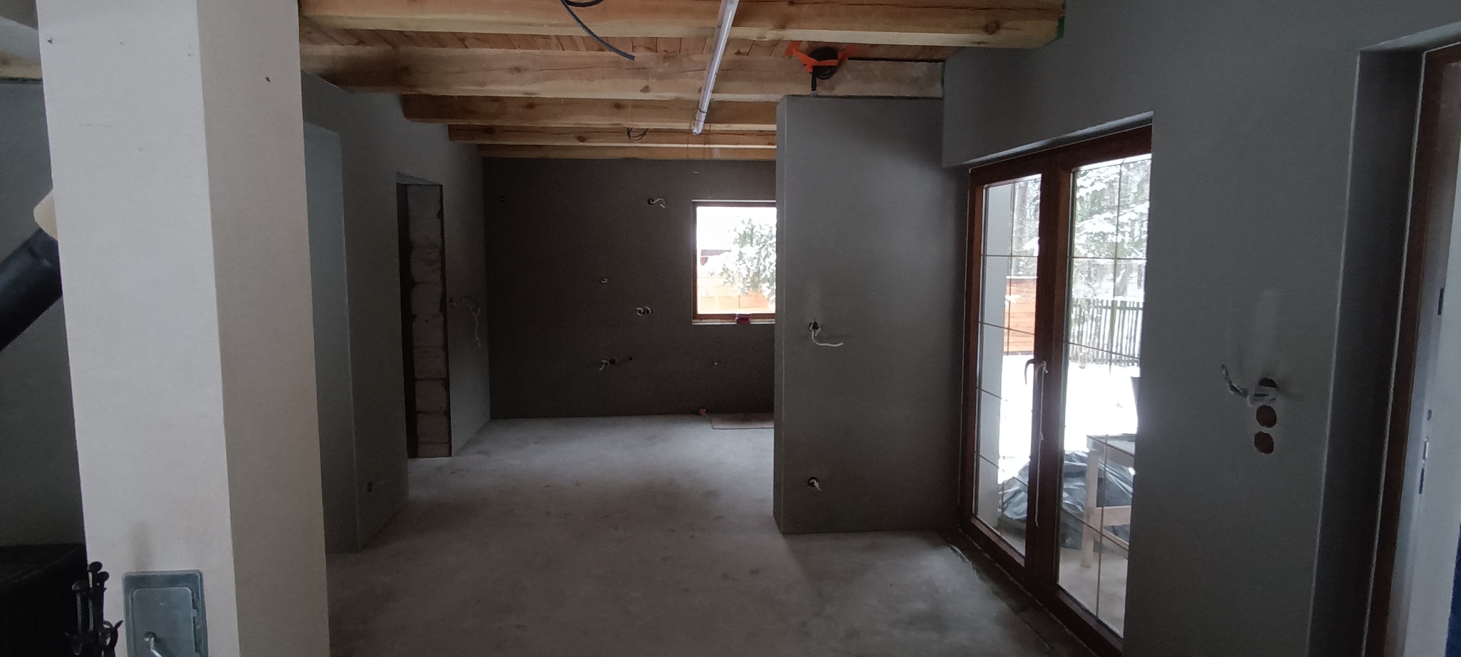 Wnętrze domu w trakcie prac wykończeniowych: ściany pokryte szarym tynkiem, widoczne instalacje elektryczne, drewniane belki stropowe, okno i drzwi balkonowe z widokiem na zimowy krajobraz.