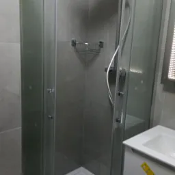 Nowoczesna kabina prysznicowa z deszczownicą i szklanymi drzwiami przesuwnymi, zamontowana w łazience z szarymi płytkami, obok umywalki z szafką.