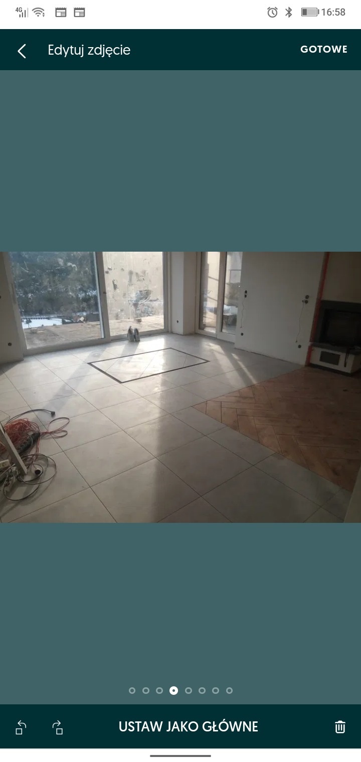 Wnętrze pokoju w trakcie remontu z częściowo ułożonymi jasnymi płytkami podłogowymi i fragmentem podłogi wyłożonym parkietem w jodełkę; widoczne przygotowania do dalszych prac wykończeniowych.