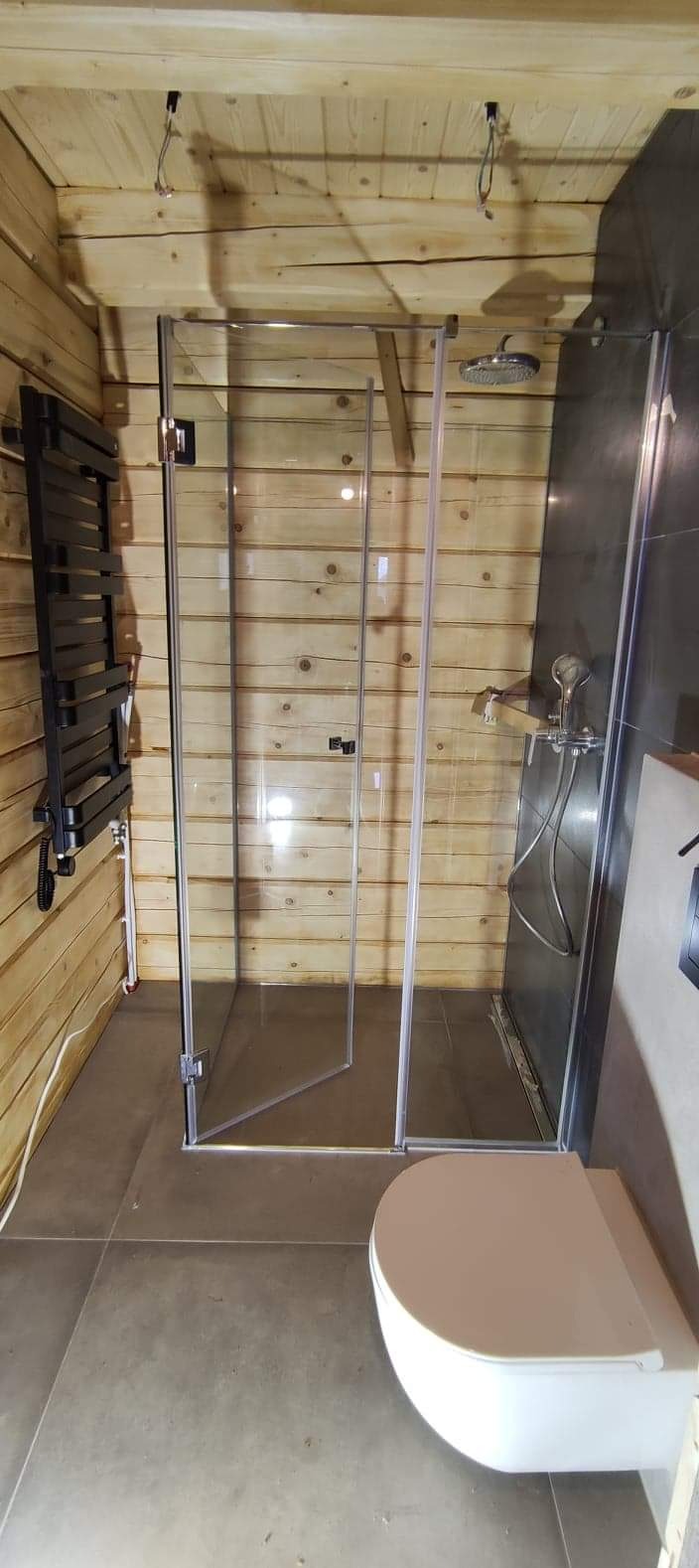 Kabina prysznicowa ze szklanymi drzwiami w łazience wykończonej drewnem i szarą płytką, widoczny grzejnik drabinkowy i wisząca toaleta.