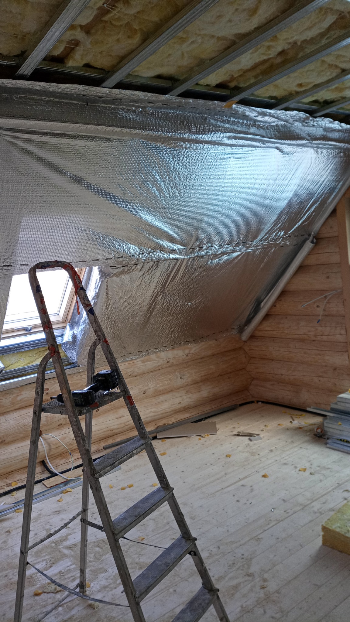 Montaż folii termoizolacyjnej na poddaszu z drewnianą boazerią, widoczna drabina i okno dachowe.