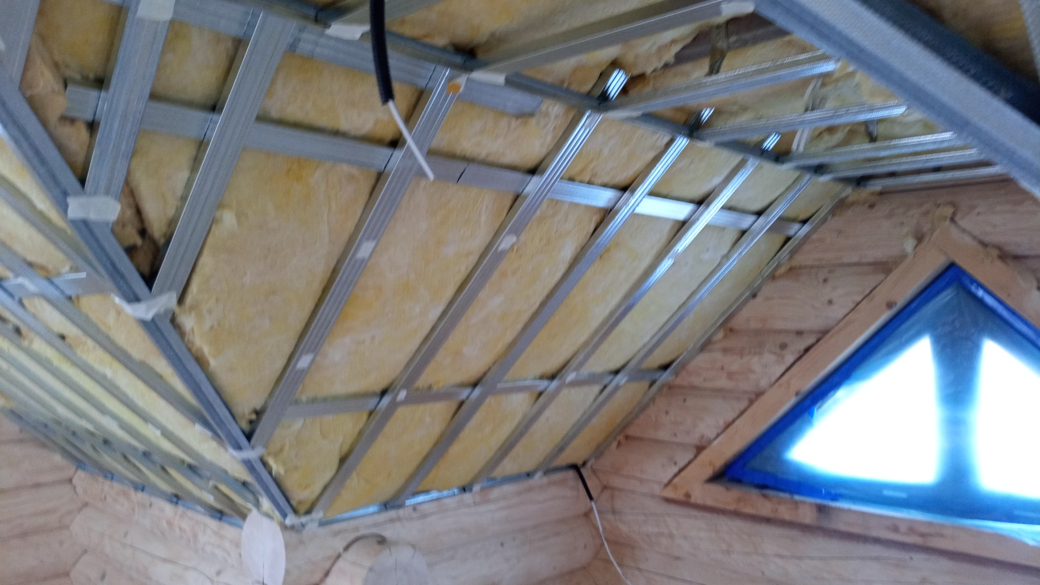 Montaż izolacji z wełny mineralnej w drewnianym domu, widoczne metalowe profile pod płyty gipsowo-kartonowe i trójkątne okno.