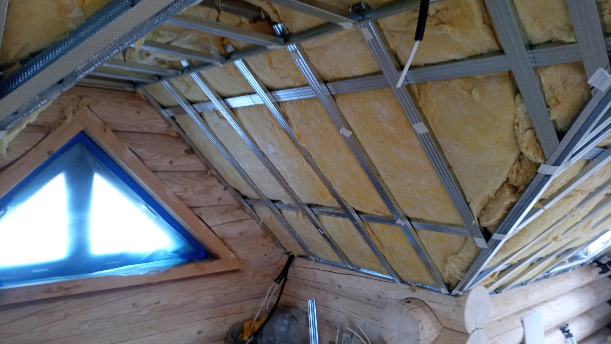 Skośny drewniany sufit z oknem, ocieplony wełną mineralną i przygotowany do montażu płyt kartonowo-gipsowych, widoczne metalowe profile.