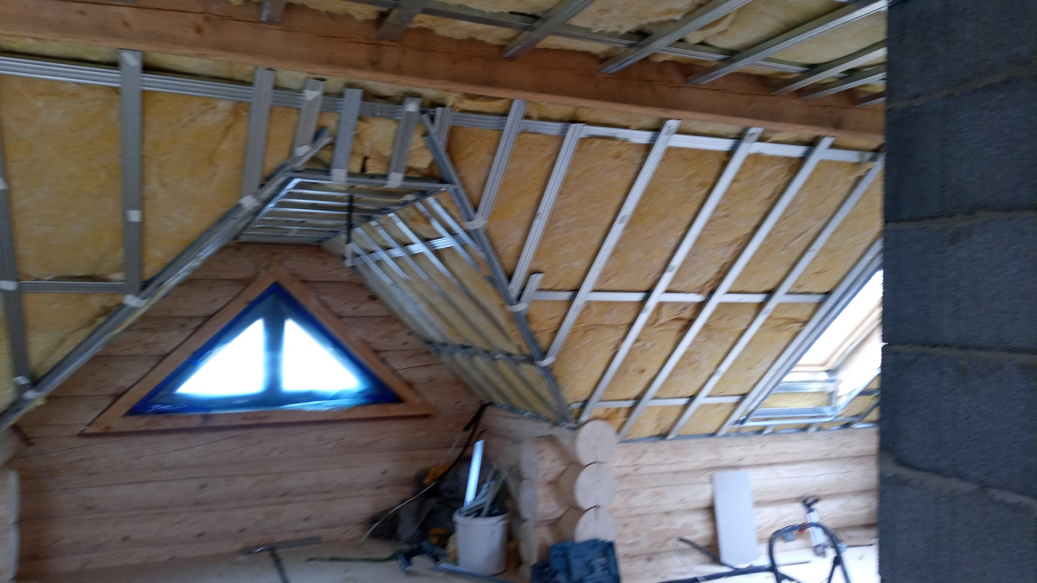 Poddasze w trakcie izolacji, widoczne wełna mineralna i metalowa konstrukcja pod płyty gipsowo-kartonowe, trójkątne okno z niebieską folią zabezpieczającą.