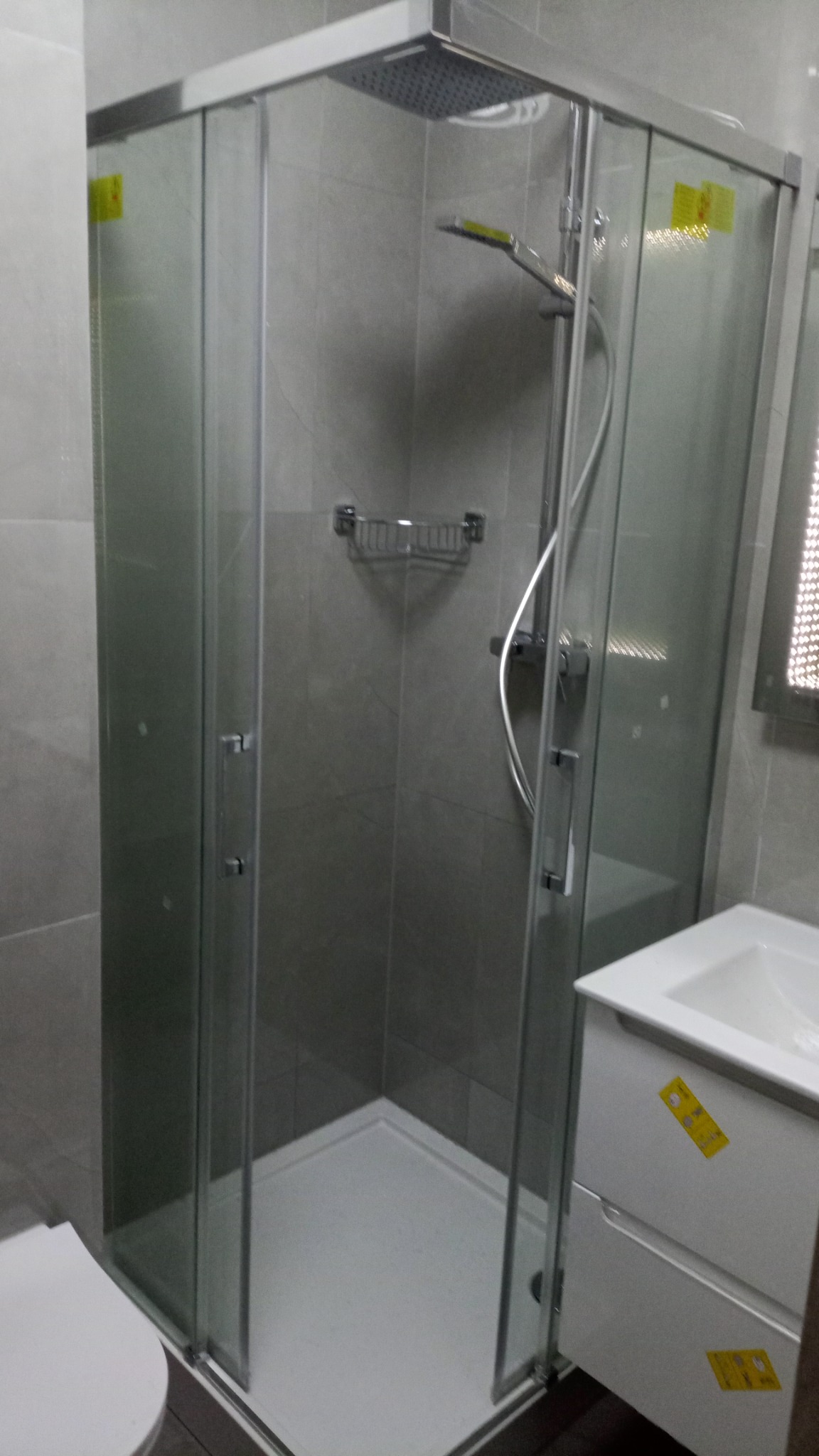 Nowoczesna kabina prysznicowa z deszczownicą i szklanymi drzwiami przesuwnymi, zamontowana w łazience z szarymi płytkami, obok umywalki z szafką.