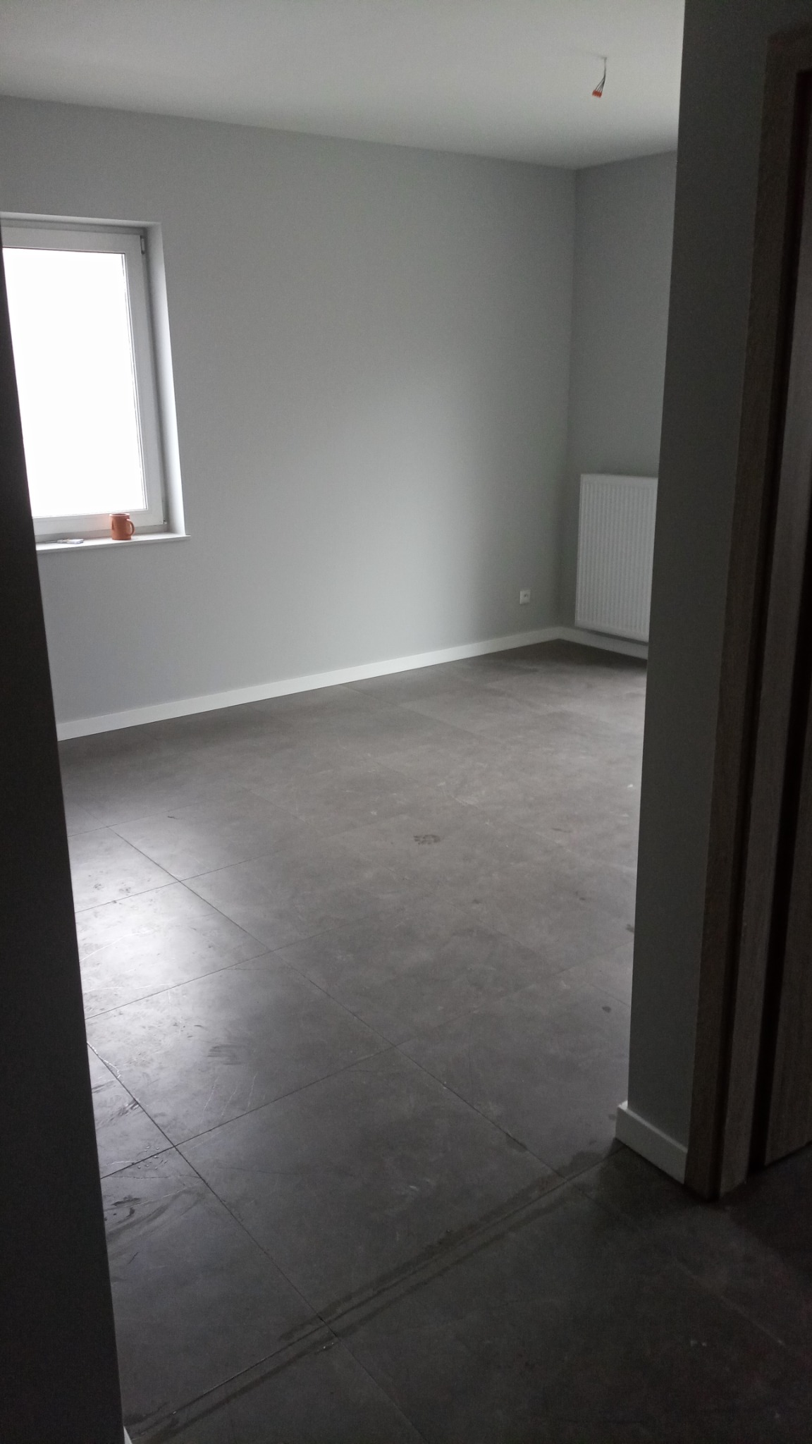 Szare płytki podłogowe w pustym, pomalowanym na szaro pokoju z grzejnikiem i kubkiem na parapecie.