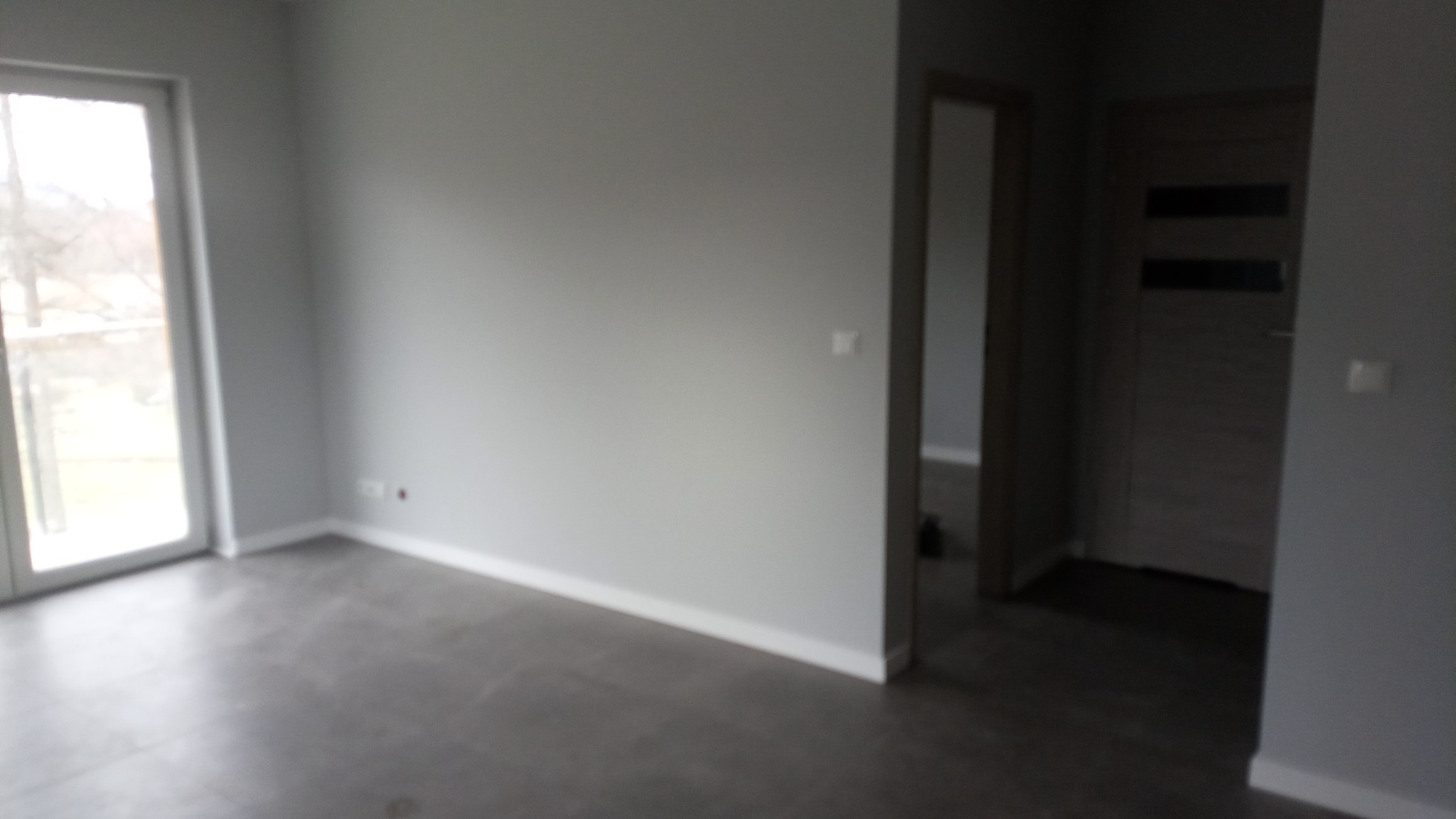 Pusty, szary pokój z szarą podłogą i białymi listwami przypodłogowymi, widoczne okno balkonowe i drzwi wewnętrzne.