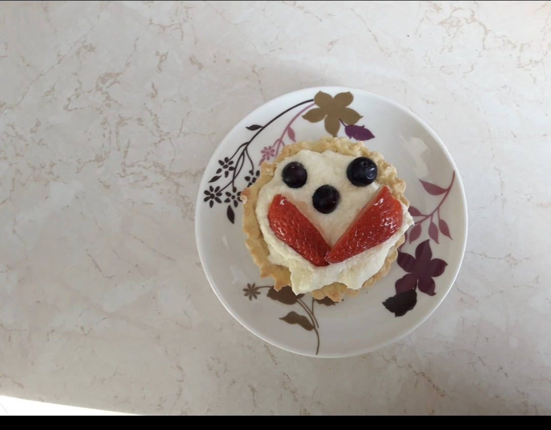 Tarta z kremem, truskawkami i borówkami ułożonymi w kształt uśmiechniętej buzi na talerzyku z kwiatowym wzorem, na blacie imitującym marmur.