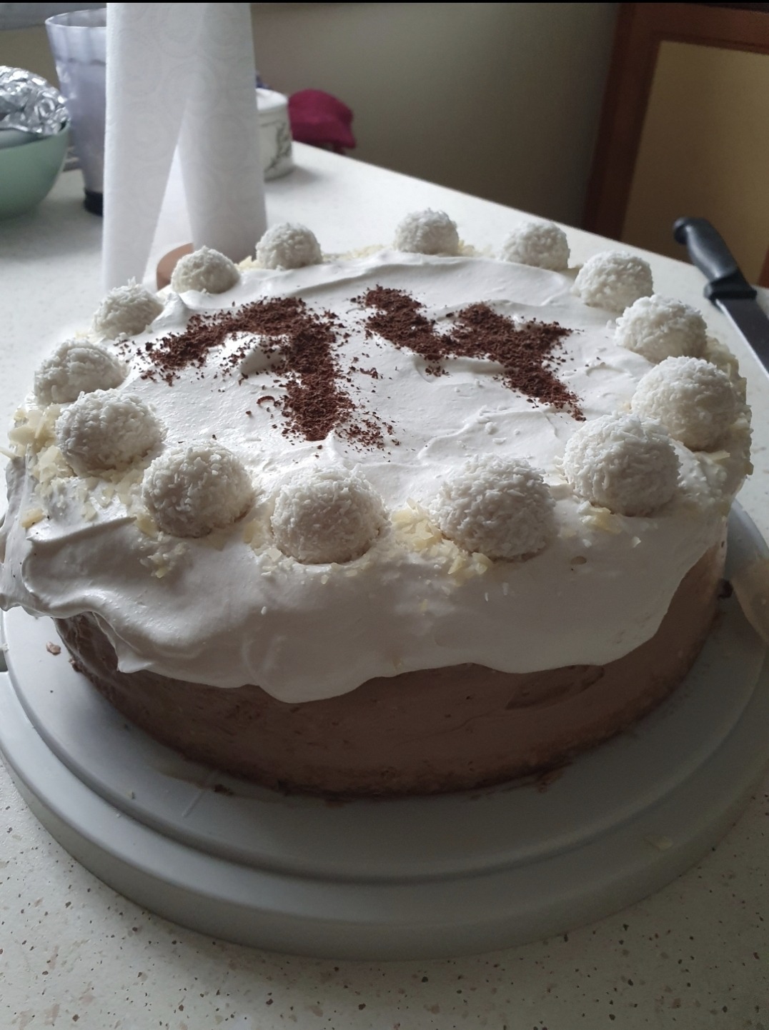 Tort czekoladowy z białym kremem, kokosowymi kulkami i posypką z ciemnej czekolady ułożoną w kształt cyfry 14.