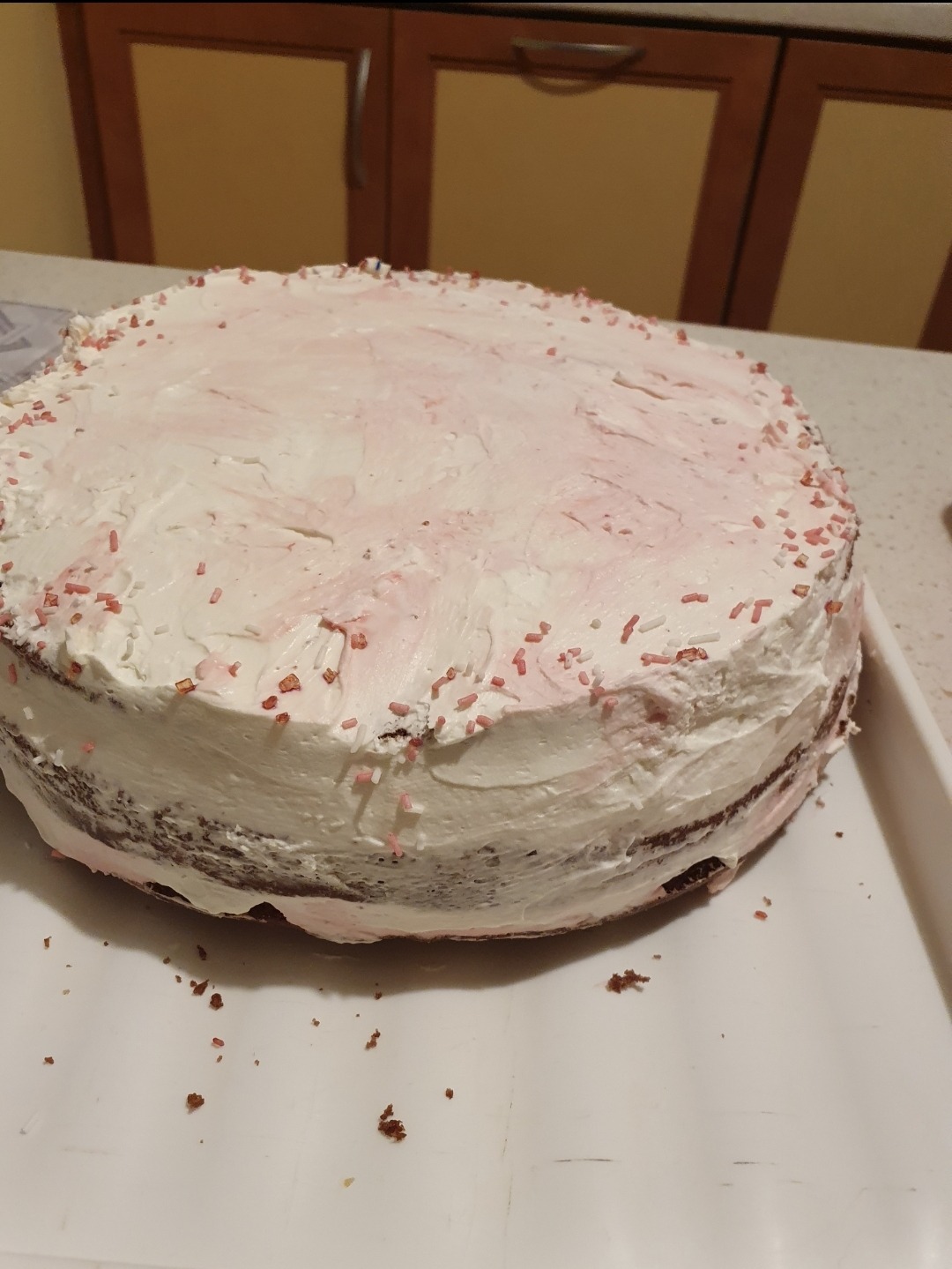Okrągły tort z kremem, posypany różowymi posypkami, na białej tacy, widoczne brązowe warstwy ciasta.