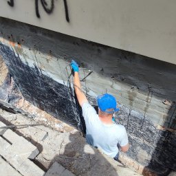 INIEKT - SYSTEM Molski Piotr - Pracownik w niebieskiej czapce i rękawicach natryskuje preparat na fundament budynku w Ostrówku, widoczne elementy izolacji oraz drabina.