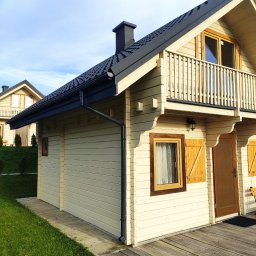 FORTIS HOUSE Budowa domów z drewna - Jasny dom szkieletowy z drewnianymi okiennicami i balkonem, widok z boku na tle zielonego trawnika i błękitnego nieba, z widocznym fragmentem drugiego domu w tle.