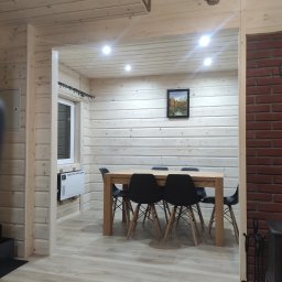 FORTIS HOUSE Budowa domów z drewna - Wnętrze domu wykończonego drewnem: jasne ściany z desek, drewniany stół otoczony czarnymi krzesłami, obraz na ścianie i grzejnik elektryczny pod oknem.