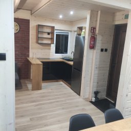 FORTIS HOUSE Budowa domów z drewna - Wnętrze domu z drewnianą konstrukcją, widoczna kuchnia z czarnymi szafkami, drewnianym blatem i ceglaną ścianą z zegarem, jasna drewniana podłoga i sufit, gaśnica na ścianie.