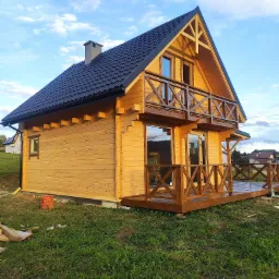 Nowoczesny, dwukondygnacyjny dom z jasnego drewna z ciemnym dachem, balkonem i tarasem, widok z boku na tle błękitnego nieba.