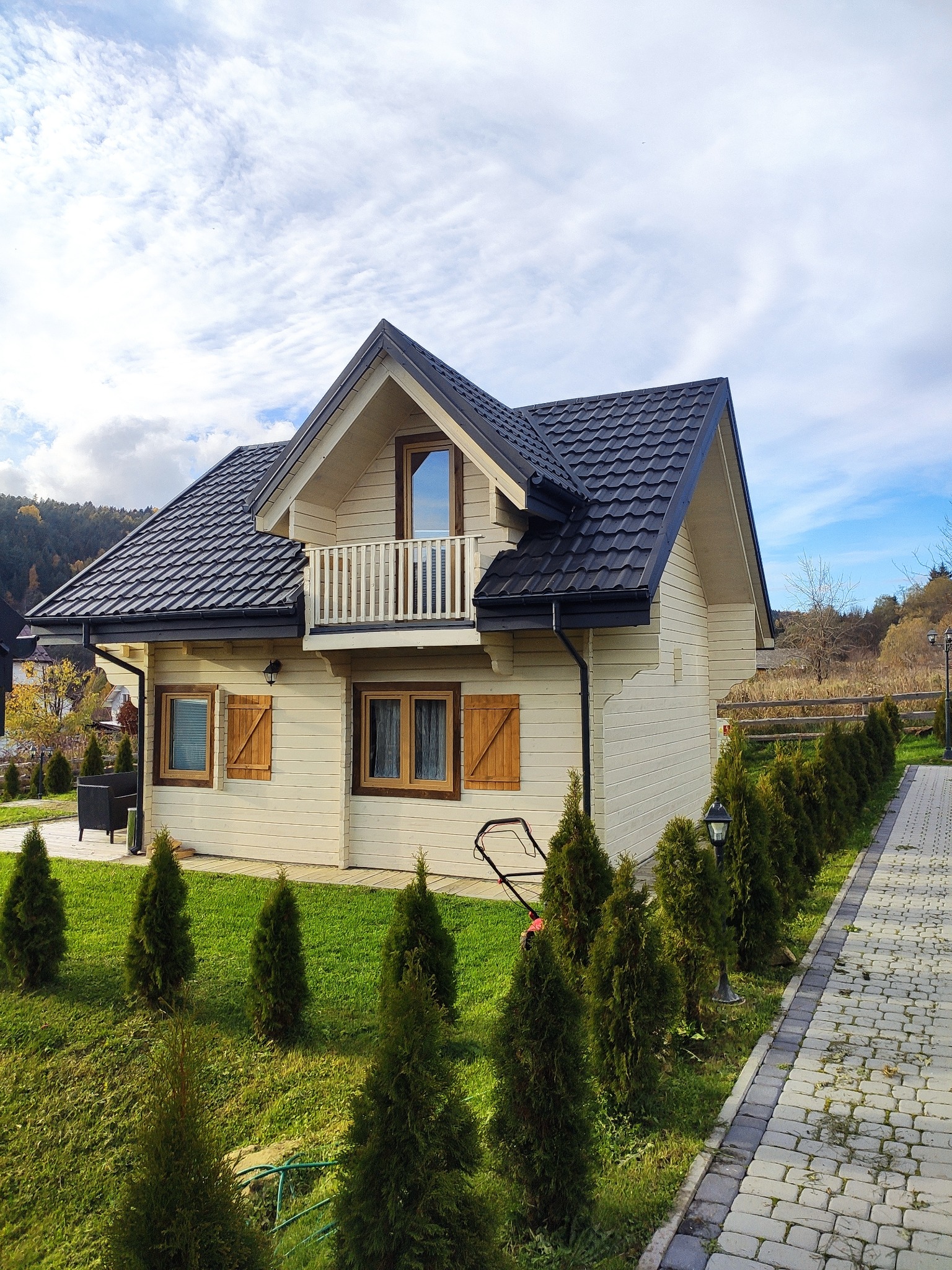 Jasny, parterowy dom z balkonem, czarnym dachem i drewnianymi okiennicami, otoczony trawnikiem z tujami, widok z boku na tle błękitnego nieba.