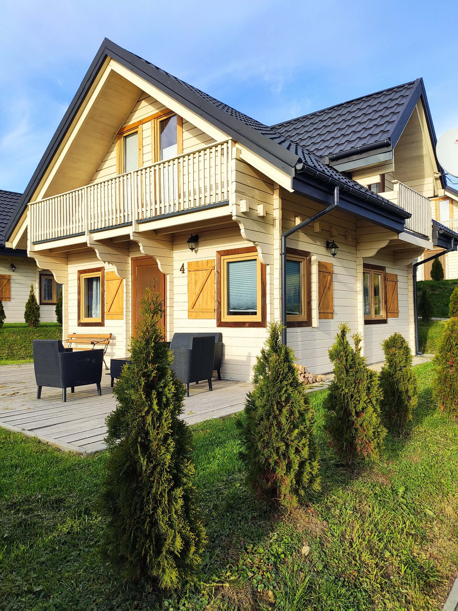 Jasny, parterowy dom z drewnianą elewacją, balkonem i ciemnym dachem, numer '4' na elewacji, drewniane okiennice, czarne meble ogrodowe na tarasie, rząd małych drzewek iglastych wzdłuż trawnika...