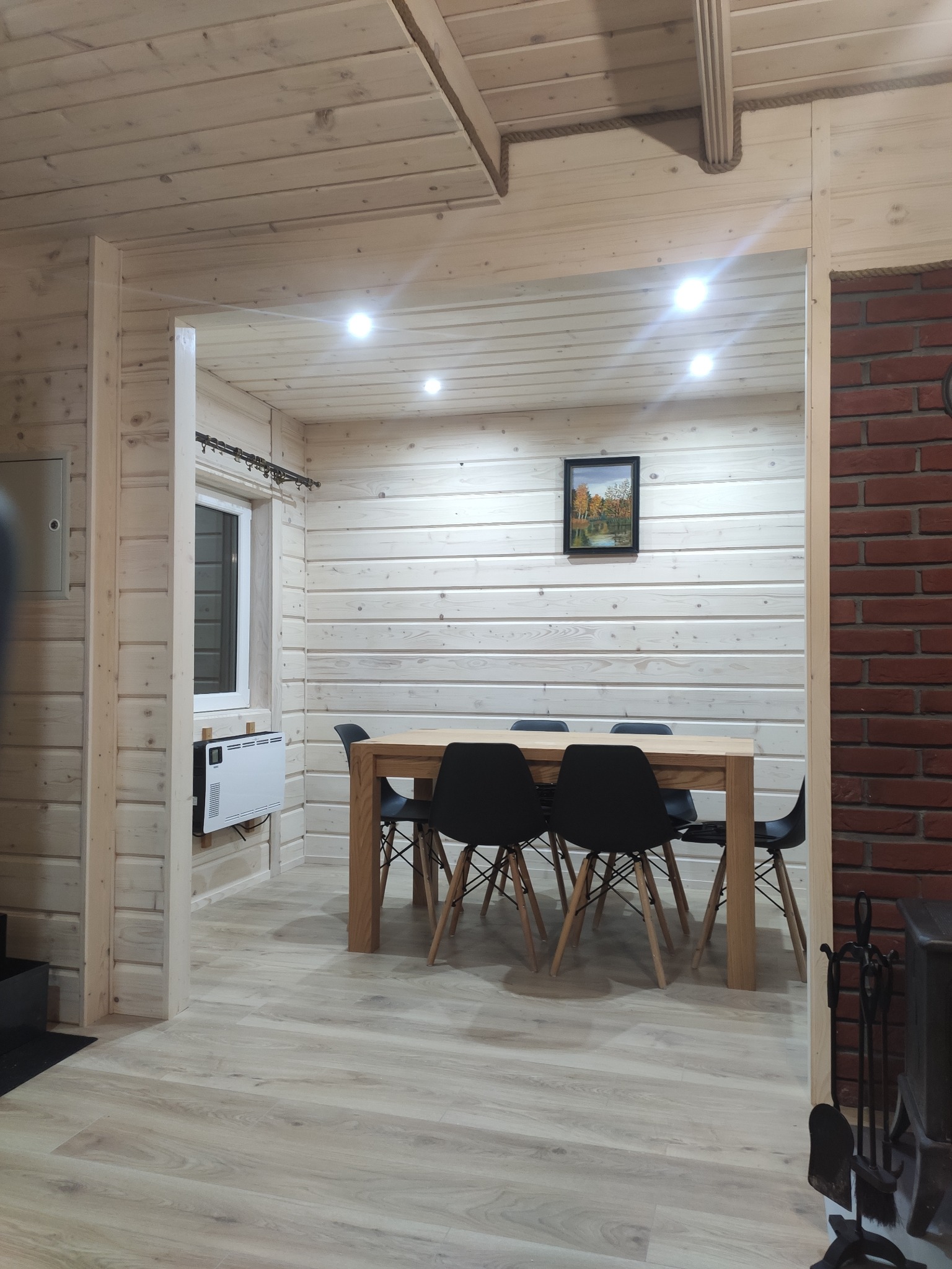 Wnętrze domu wykończonego drewnem: jasne ściany z desek, drewniany stół otoczony czarnymi krzesłami, obraz na ścianie i grzejnik elektryczny pod oknem.
