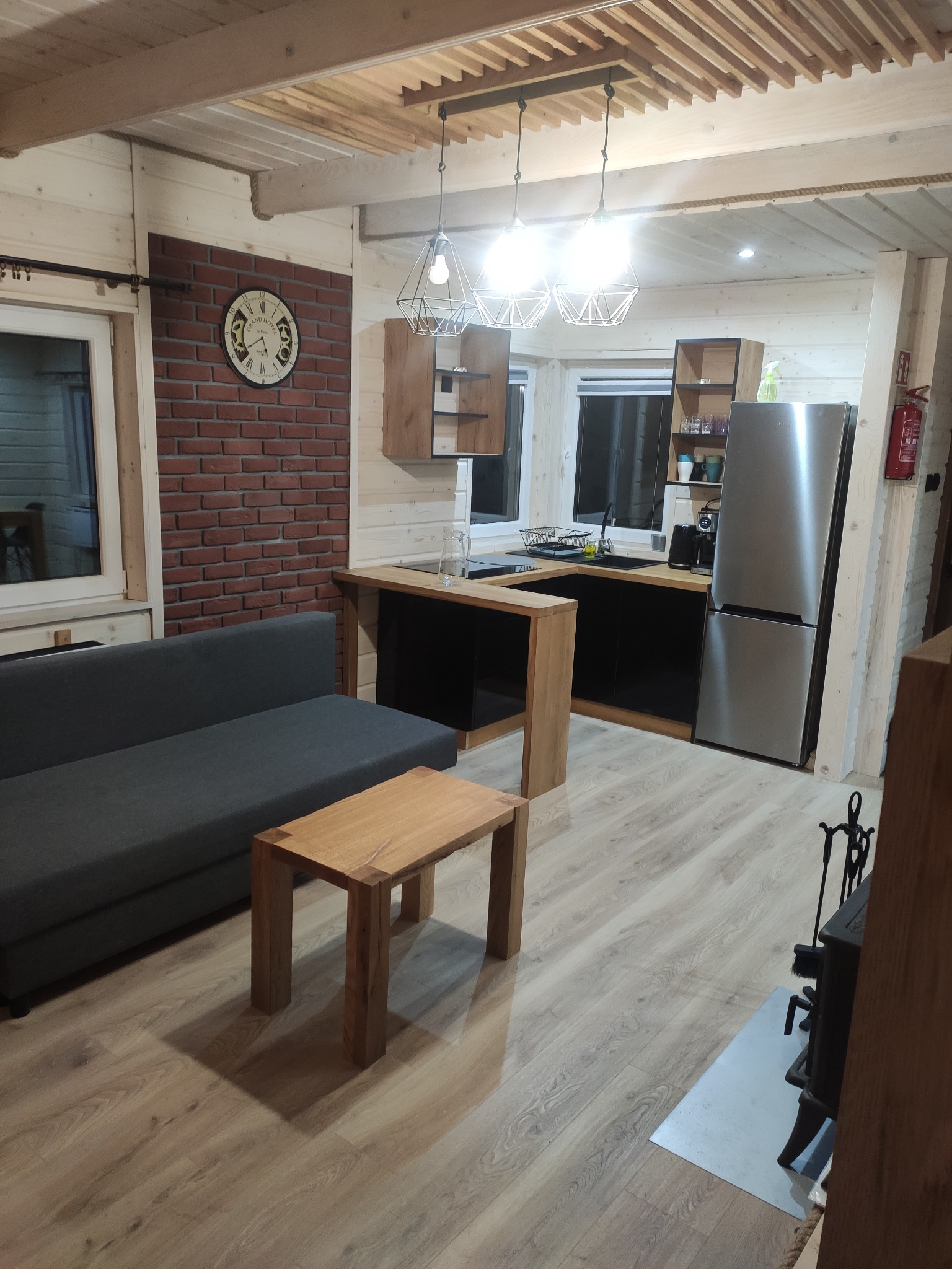 Jasne wnętrze domu z drewnianymi ścianami, szarą sofą, drewnianym stolikiem kawowym i aneksem kuchennym z czarnymi frontami szafek.