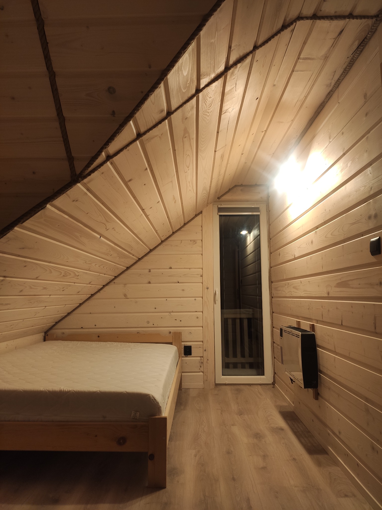 Wnętrze pokoju na poddaszu wykończonego drewnianymi panelami, z łóżkiem, oknem balkonowym i grzejnikiem elektrycznym.