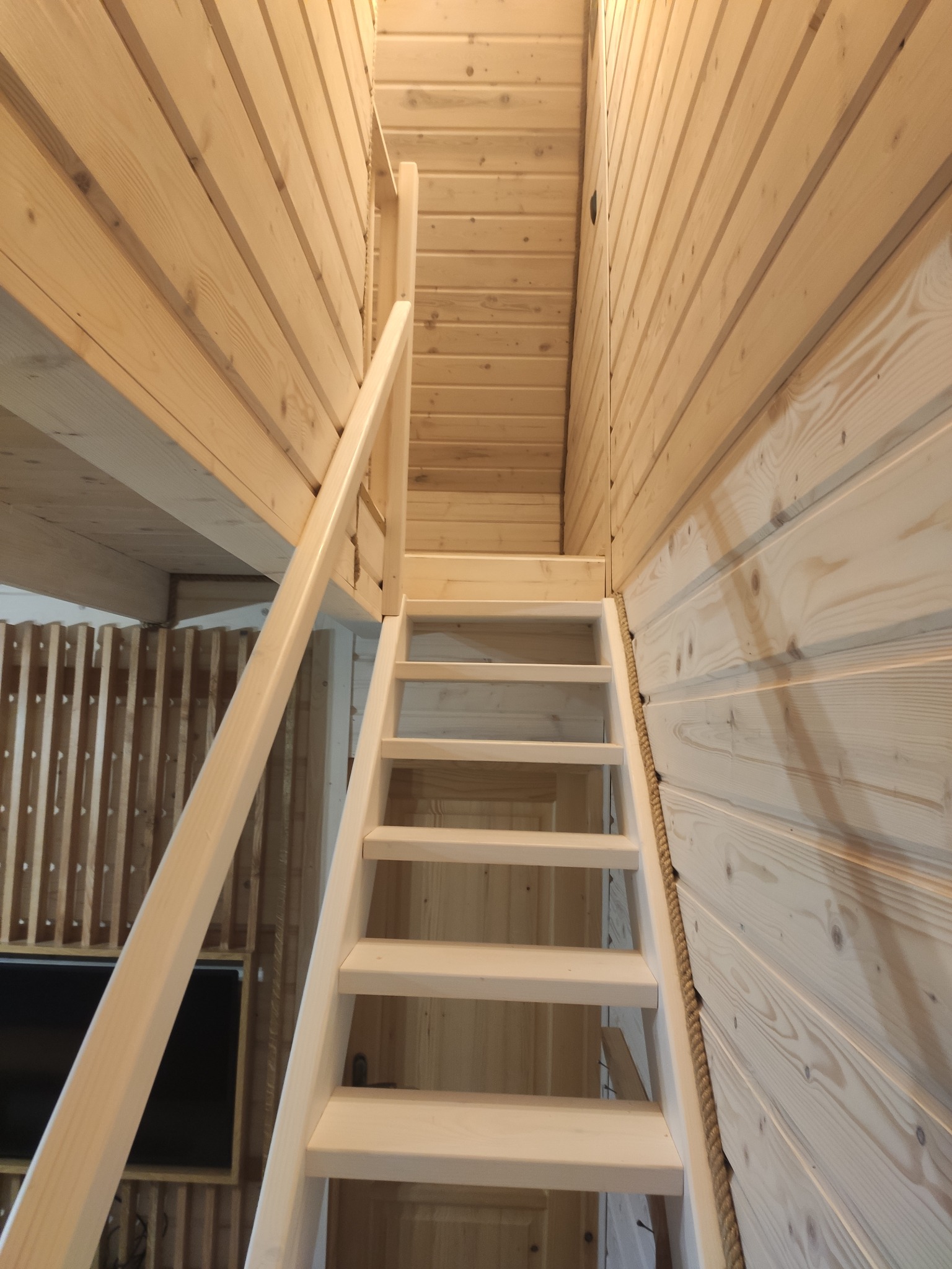 Jasne drewniane schody prowadzące na antresolę w domu wykończonym boazerią, z liną jutową jako elementem dekoracyjnym.