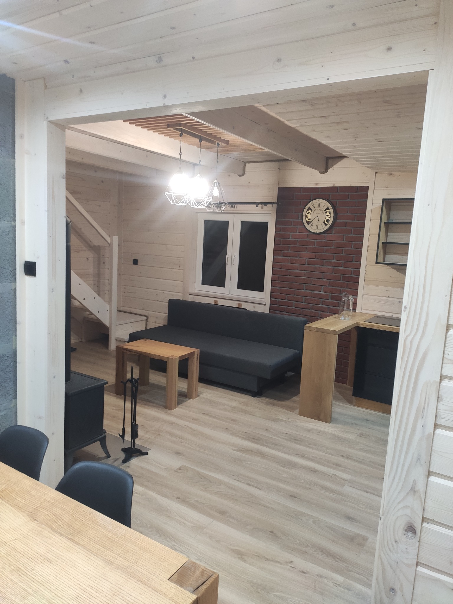 Jasne wnętrze domu z drewnianymi ścianami, widok z jadalni na salon z kominkiem, schody i aneks kuchenny z ceglaną ścianą.