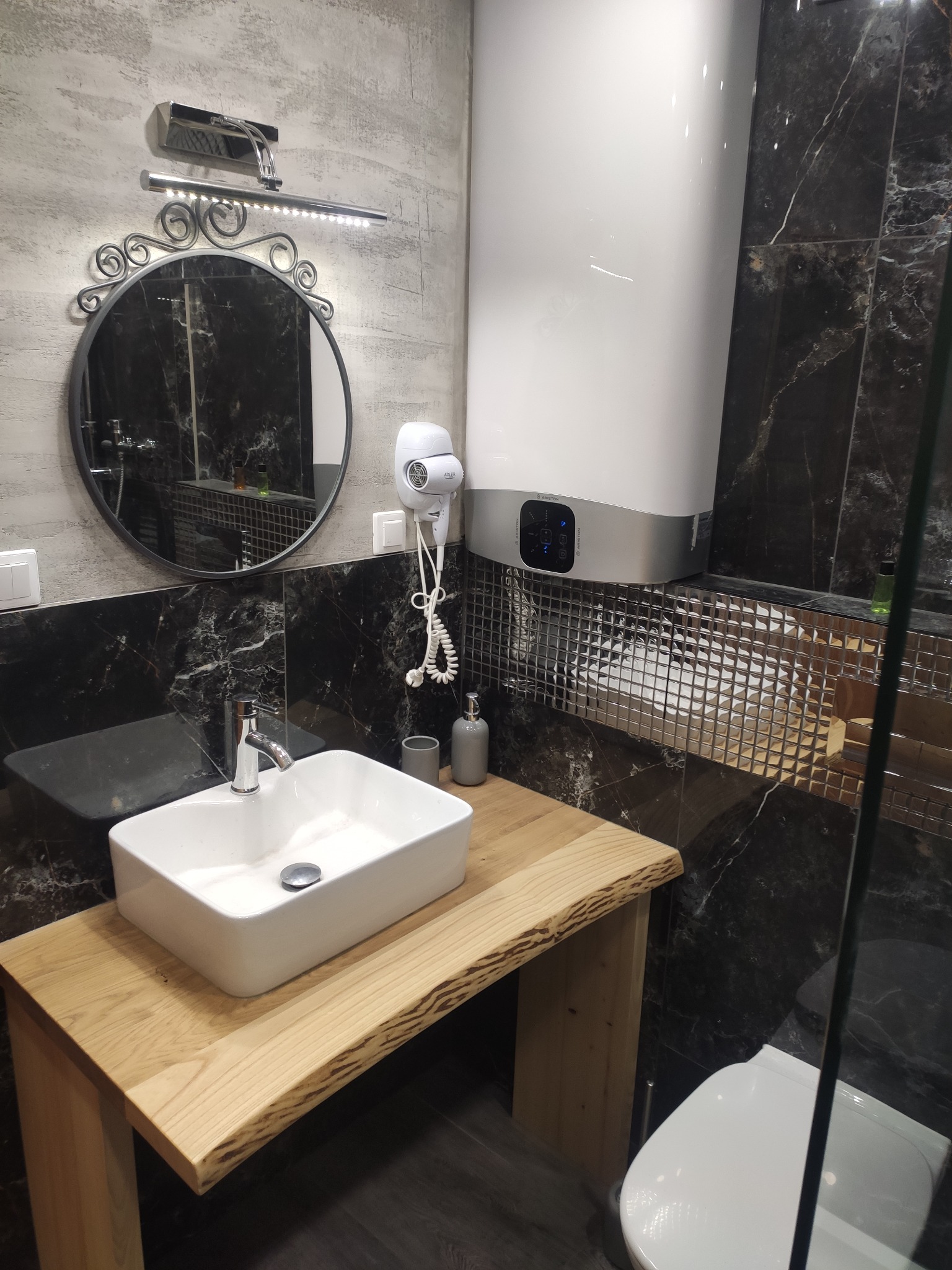 Nowoczesna łazienka z umywalką nablatową na drewnianym blacie, okrągłym lustrem z ozdobnym oświetleniem, grzejnikiem elektrycznym i czarnymi marmurkowymi płytkami.