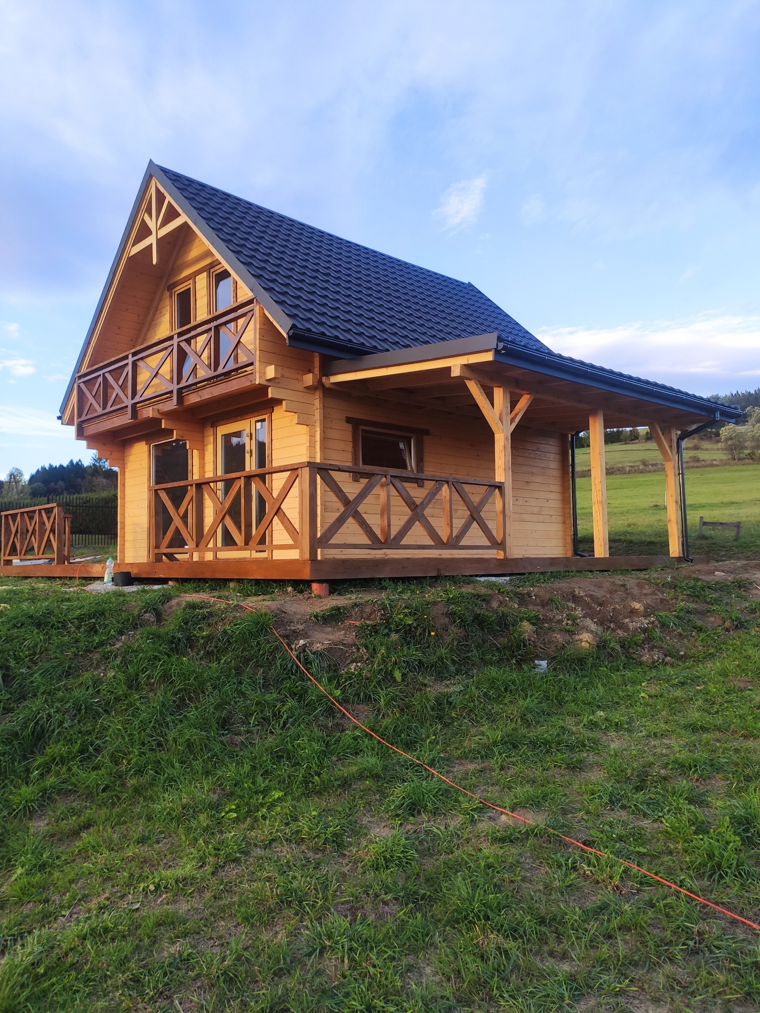 Nowoczesny, parterowy dom z drewna z balkonem, wykończony czarną dachówką, na tle zielonego wzgórza i błękitnego nieba.