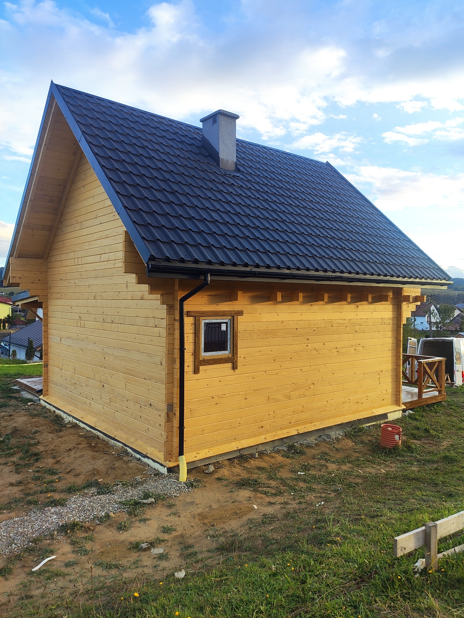 Zewnętrzny widok nowego, jasnego, drewnianego domu z ciemnym dachem, małym oknem i małym tarasem, położonego na wzgórzu z widokiem na inne domy w oddali.