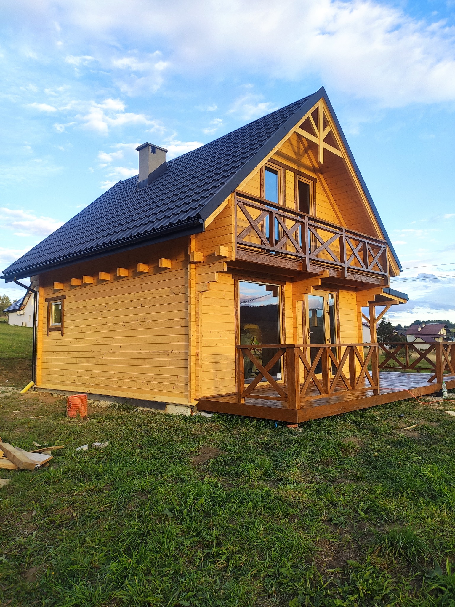 Nowoczesny, dwukondygnacyjny dom z jasnego drewna z ciemnym dachem, balkonem i tarasem, widok z boku na tle błękitnego nieba.