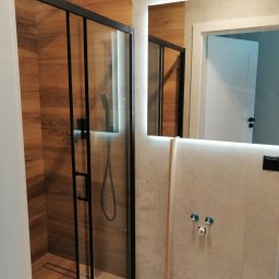 Usługi remontowo-wykończeniowe Matysiak Mariusz - Nowoczesna kabina prysznicowa z czarnymi profilami i drewnopodobnymi płytkami, oświetlone lustro z widocznym fragmentem drzwi do innego pomieszczenia.