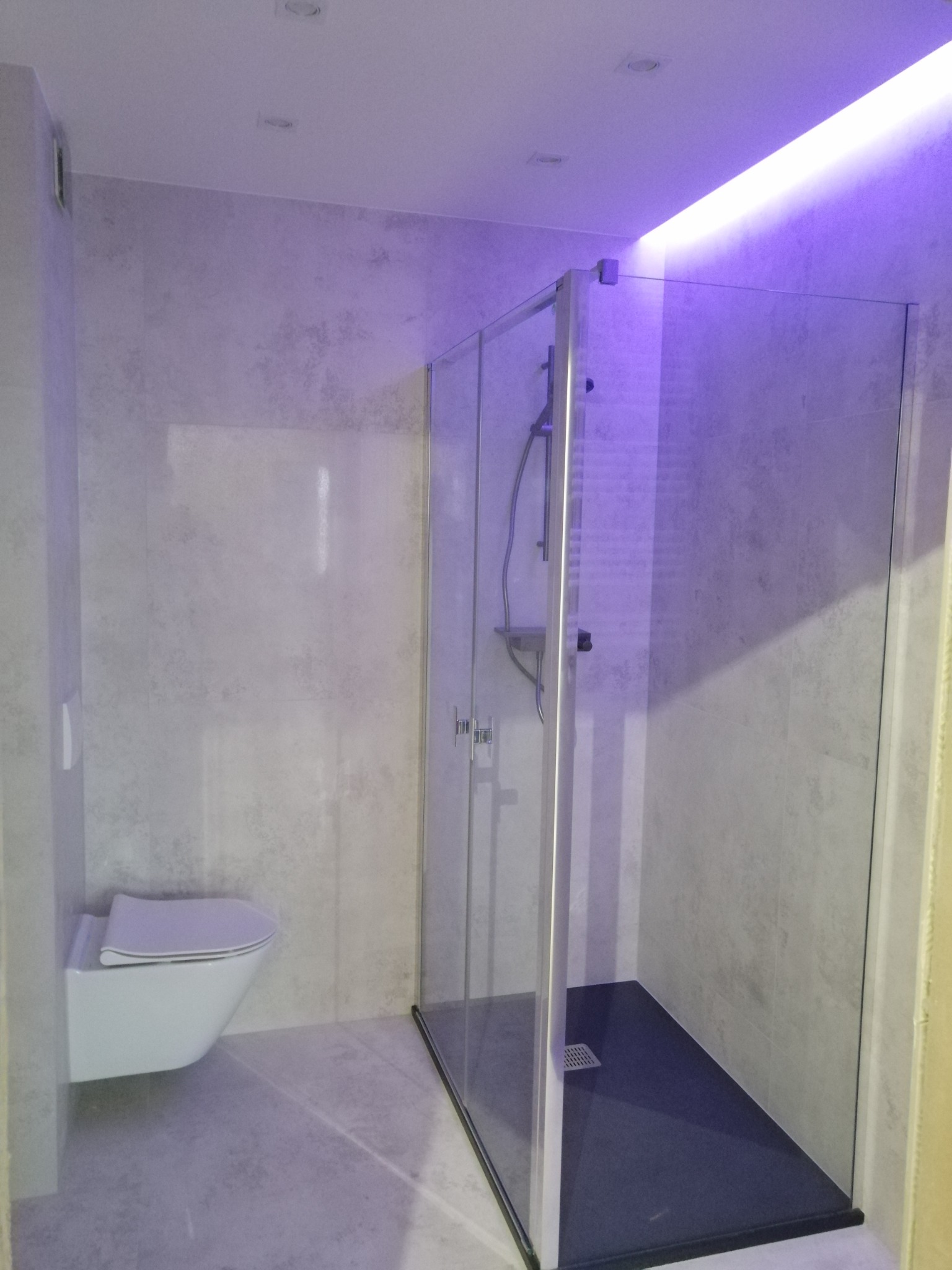 Nowoczesna łazienka z kabiną prysznicową typu walk-in z czarnym brodzikiem i podświetleniem LED w odcieniach fioletu, wisząca miska WC.