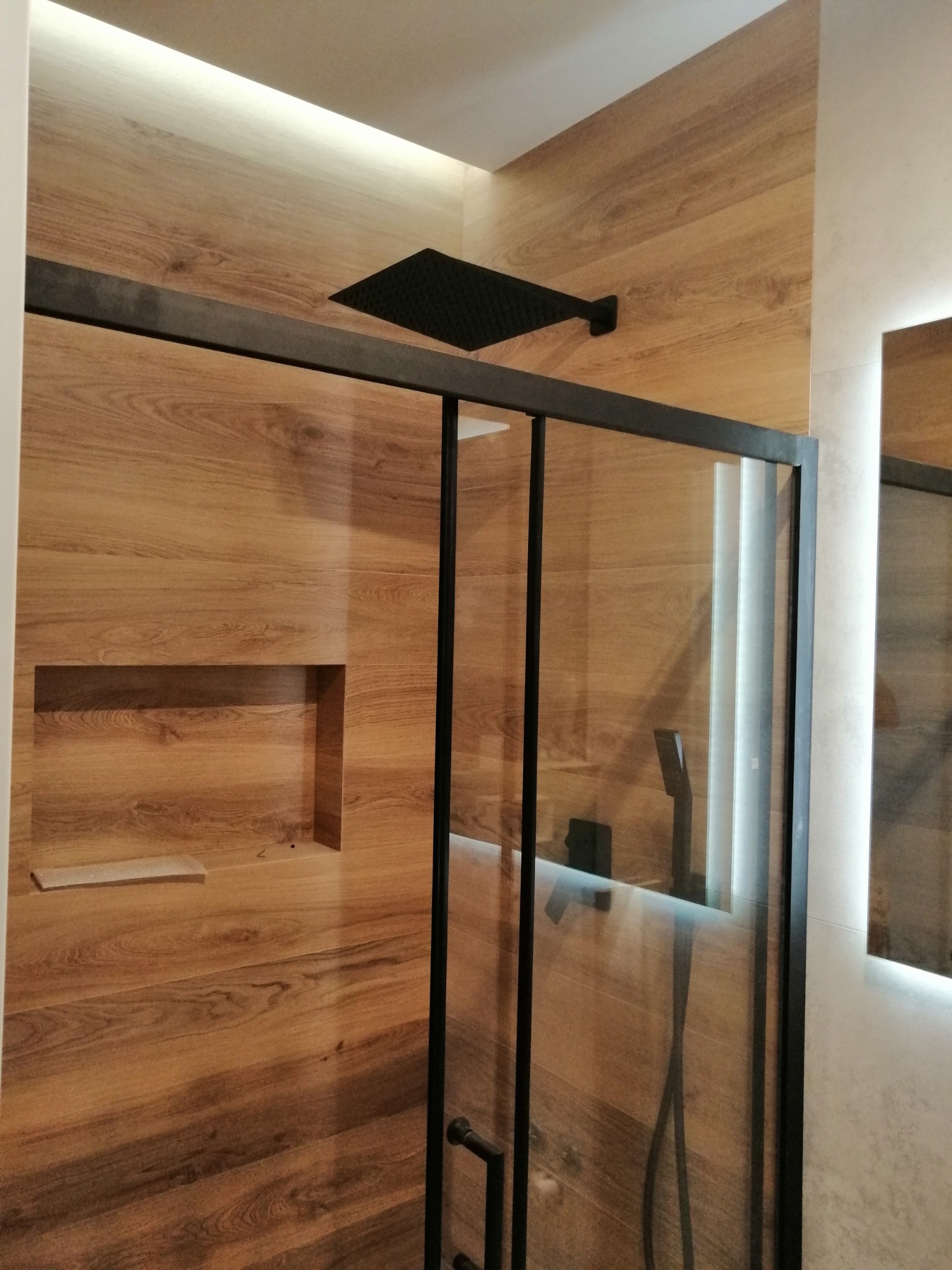 Nowoczesna kabina prysznicowa z czarną armaturą i deszczownicą, drewnopodobne płytki na ścianach z wnęką, oświetlenie LED podsufitowe i lustro z podświetleniem.