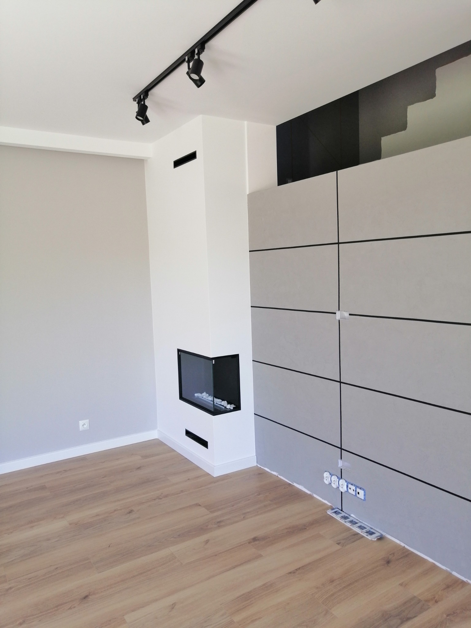 Nowoczesne wnętrze z drewnianą podłogą, ścianą z paneli i minimalistycznym kominkiem narożnym z czarną obudową, oświetlenie szynowe na suficie, widoczne gniazdka elektryczne w ścianie.