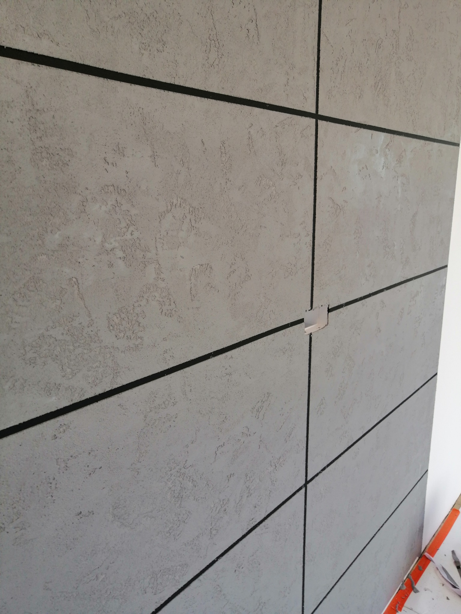 Wykończona ściana z szarego tynku strukturalnego podzielona czarnymi liniami, widoczna puszka elektryczna i fragment narzędzi malarskich w prawym dolnym rogu.